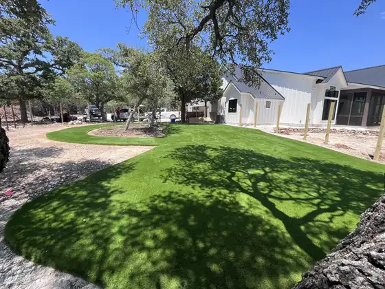 Artificial turf lawn in a backyard beside a white home, shaded by oak trees